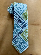 Load image into Gallery viewer, A patterned necktie with a light blue base and geometric tribal-style prints in navy blue and olive green, displayed on a brown wooden surface.