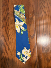 Load image into Gallery viewer, A blue necktie with a tropical floral print featuring white flowers and green leaves, laid flat on a brown wooden surface.