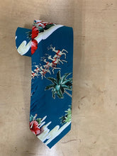 Load image into Gallery viewer, A blue Hawaiian-style necktie with a tropical print featuring palm trees, hibiscus flowers, and Santa Claus in sleighs, displayed on a light wood background.