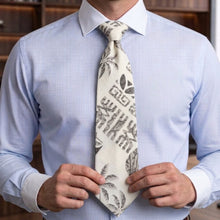 Load image into Gallery viewer, A man wearing a light blue dress shirt and a cream-colored tie with a black Hawaiian-style graphic pattern, adjusting the tie with both hands.