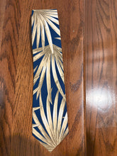 Load image into Gallery viewer, A necktie with a navy blue background and a metallic gold palm leaf pattern, laid flat on a dark wood surface.