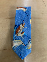 Load image into Gallery viewer, A blue necktie with a pattern of sailfish and marlin fish, laid flat on a light brown wooden surface.