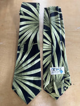 Load image into Gallery viewer, A men's necktie with a green palm leaf pattern on a black background, laid flat on a light wood surface next to its white paper tag.
