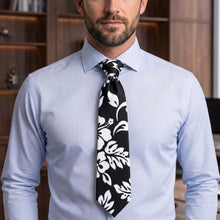 Load image into Gallery viewer, A man wearing a light blue dress shirt and a black necktie with a white floral pattern, shown from the chest up in an indoor setting with a blurred wooden background.
