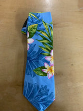 Load image into Gallery viewer, A tropical print necktie featuring blue palm leaves and pink and yellow plumeria flowers on a light blue background, displayed on a wooden surface.
