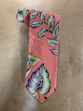 Load image into Gallery viewer, A coral pink necktie with a tropical leaf pattern in green, purple, and white, laid flat on a light brown wooden surface.
