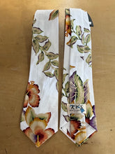 Load image into Gallery viewer, Two white neckties with a tropical floral and leaf print, laid flat on a tan wooden surface. One tie is oriented vertically, and the other is laid horizontally next to it, showing a brand label that reads 'KYS BOSTONIAN MADE NECKLACES'.
