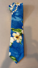 Load image into Gallery viewer, A blue necktie with a tropical floral print featuring white hibiscus flowers and green leaves, laid flat on a neutral beige surface.
