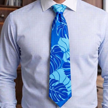Load image into Gallery viewer, A men's necktie with a tropical leaf pattern in shades of blue, worn with a light blue pinstriped dress shirt.
