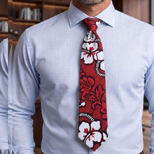Load image into Gallery viewer, A men's necktie with a red, white, and black hibiscus floral pattern, styled on a light blue striped dress shirt. The photo is cropped from the chest up, set against a blurred background of a wooden bookshelf.
